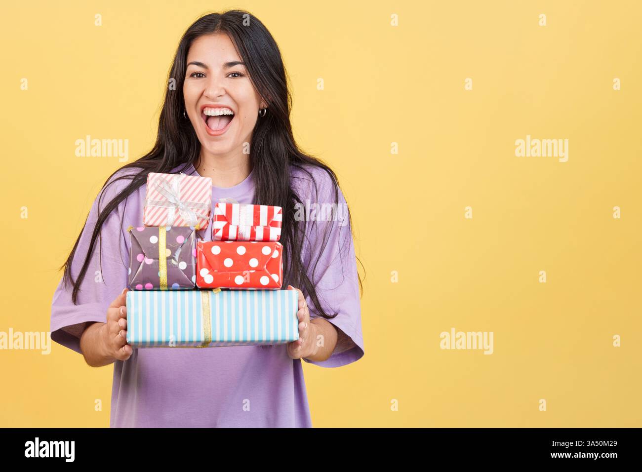 Fröhliche weiße Frau hält einen Stapel bunter Geschenke in einem hellen Atelier vor gelbem Hintergrund. Diese festliche Szene ist ideal für Feiertags- und Weihnachtskampagnen, Geschenke und Werbematerial. Stockfoto