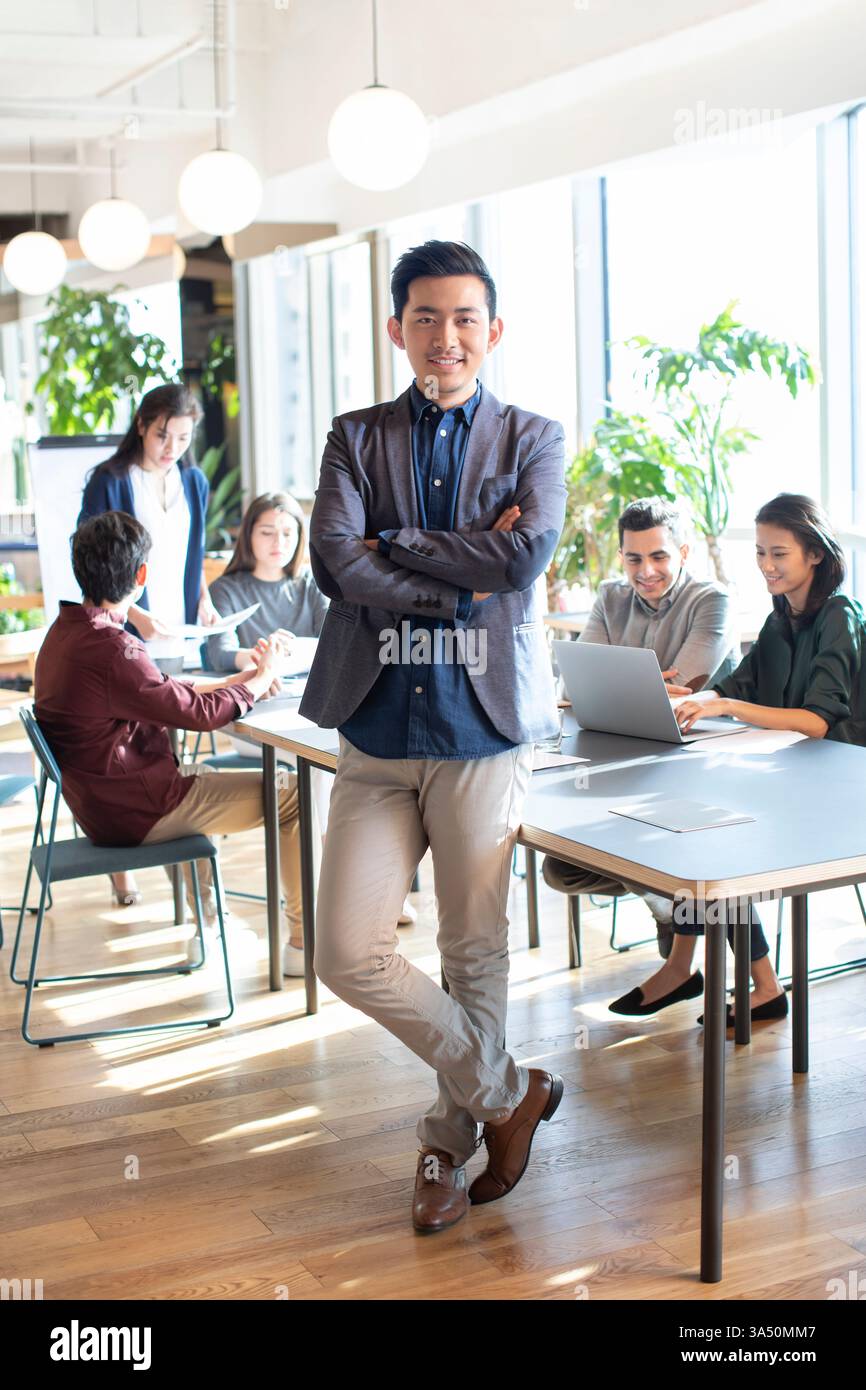 Lächelnder chinesischer Geschäftsmann in grauem Anzug, der mit überkreuzten Armen vor seinen Kollegen im Büro steht Stockfoto