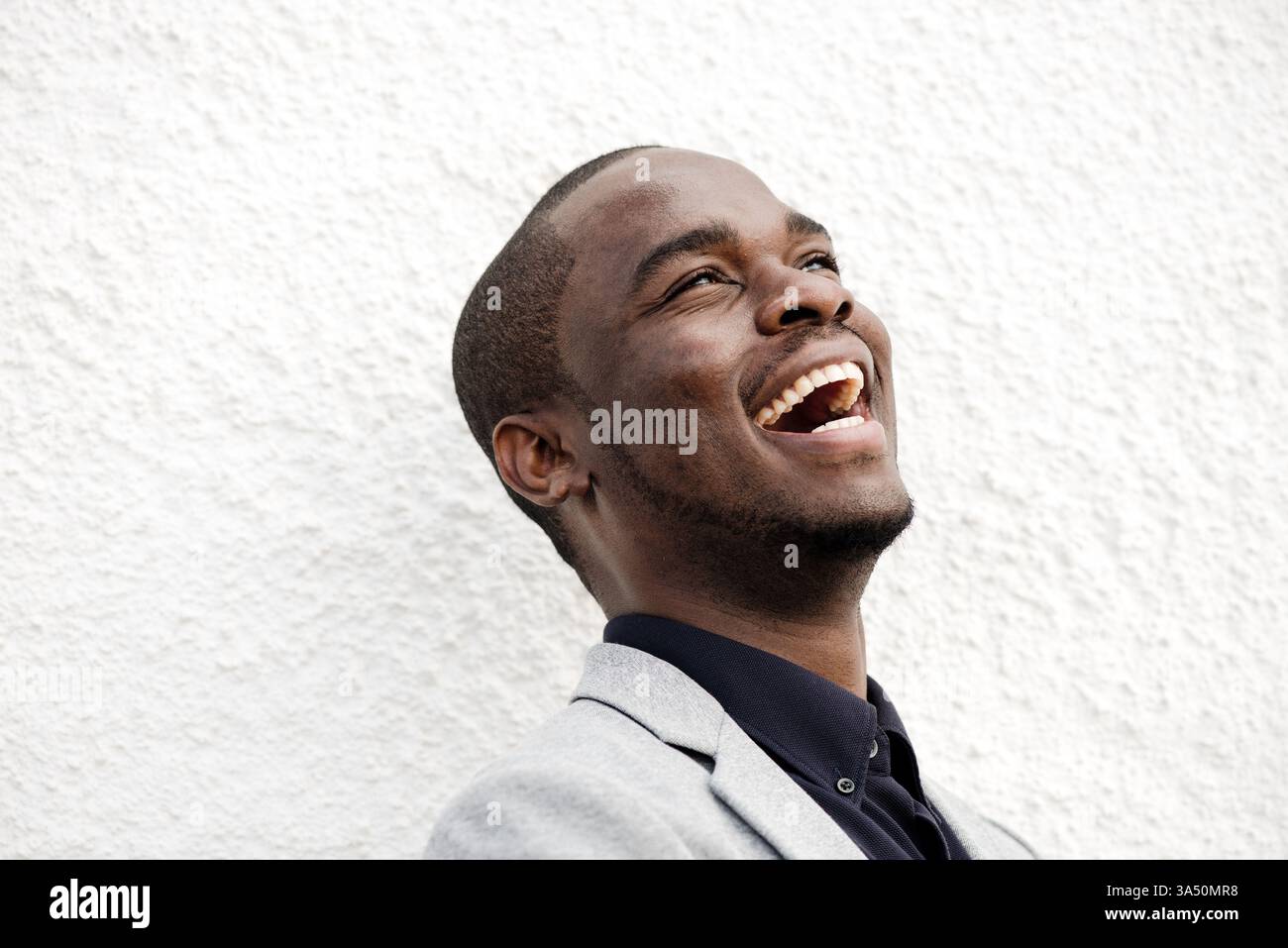Nahaufnahme Porträt eines fröhlichen Afroamerikaners, der an einer weißen Wand lacht. Das Image projiziert Selbstvertrauen und Positivität, die sich für geschäftliche und Lifestyle-Themen eignen. Ideal für Branding- oder Marketingmaterialien mit verschiedenen Profis und modernem Stil. Stockfoto