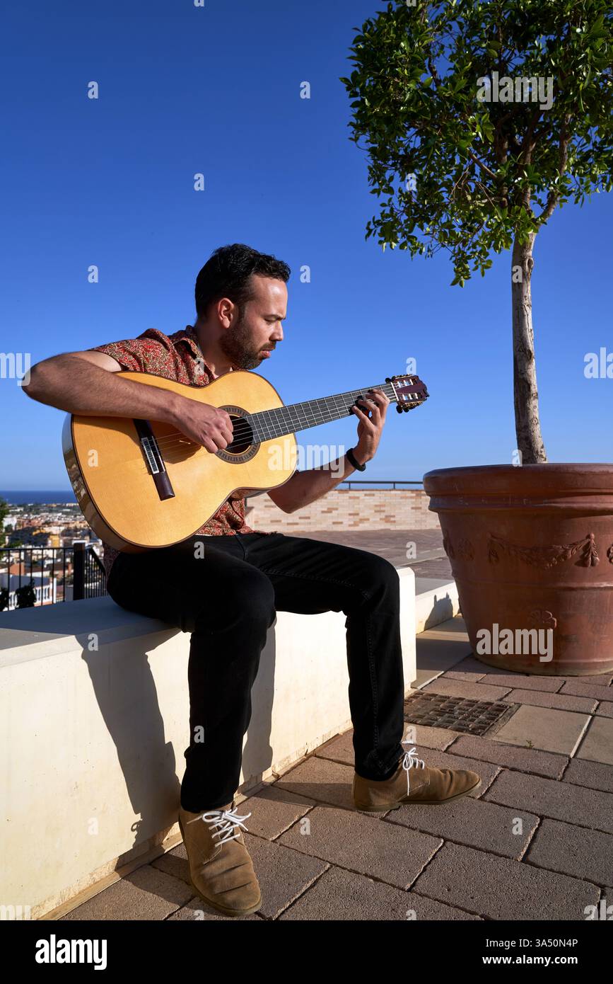 Ganzkörperporträt eines bärtigen Musikers, der draußen in der Stadt Akustikgitarre spielt. Vor blauem Himmel sitzt er am Rande einer Grenze und liefert eine melodische Straßenaufführung. Stockfoto