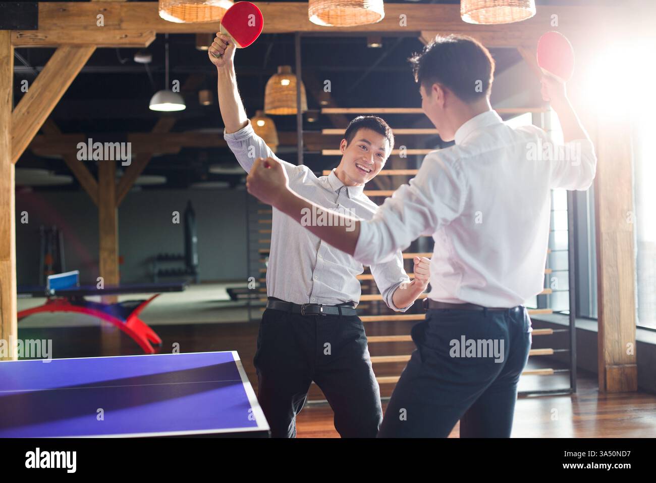 Junge chinesische Geschäftsleute spielen Tischtennis in einem modernen Büro. Der energiegeladene Moment mit Anfeuern und Teamwork macht dieses Image für Corporate Branding, Bürokultur und sportliche Kampagnen geeignet. Stockfoto