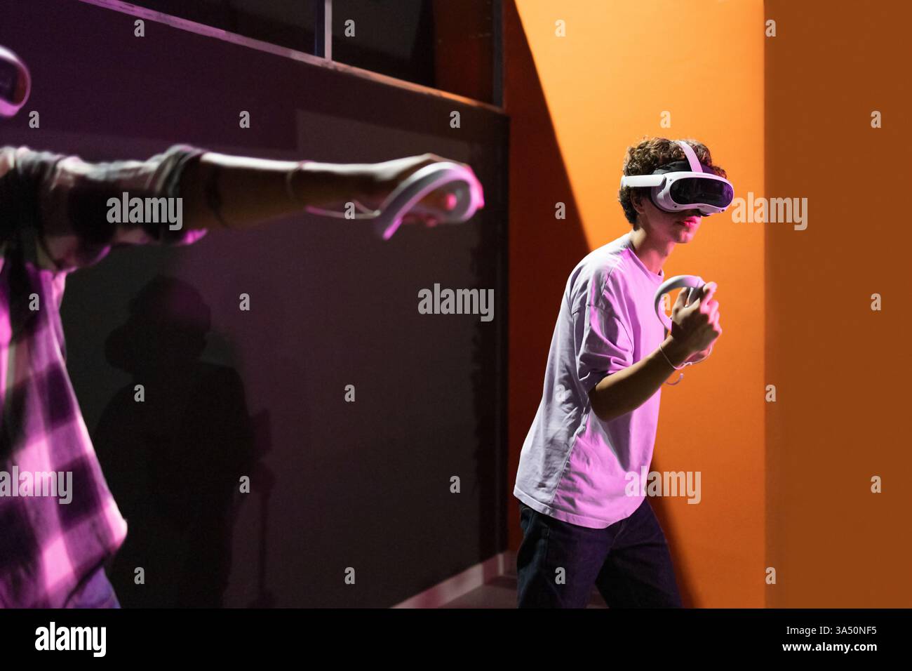 Seitenansicht von zwei Freunden in einem Spielbereich, die mit VR-Headsets und Motion-Controllern vollständig in die virtuelle Realität eingetaucht sind. Neonlichter und High-Tech-Ausrüstung schaffen eine futuristische, immersive Unterhaltungsszene. Stockfoto