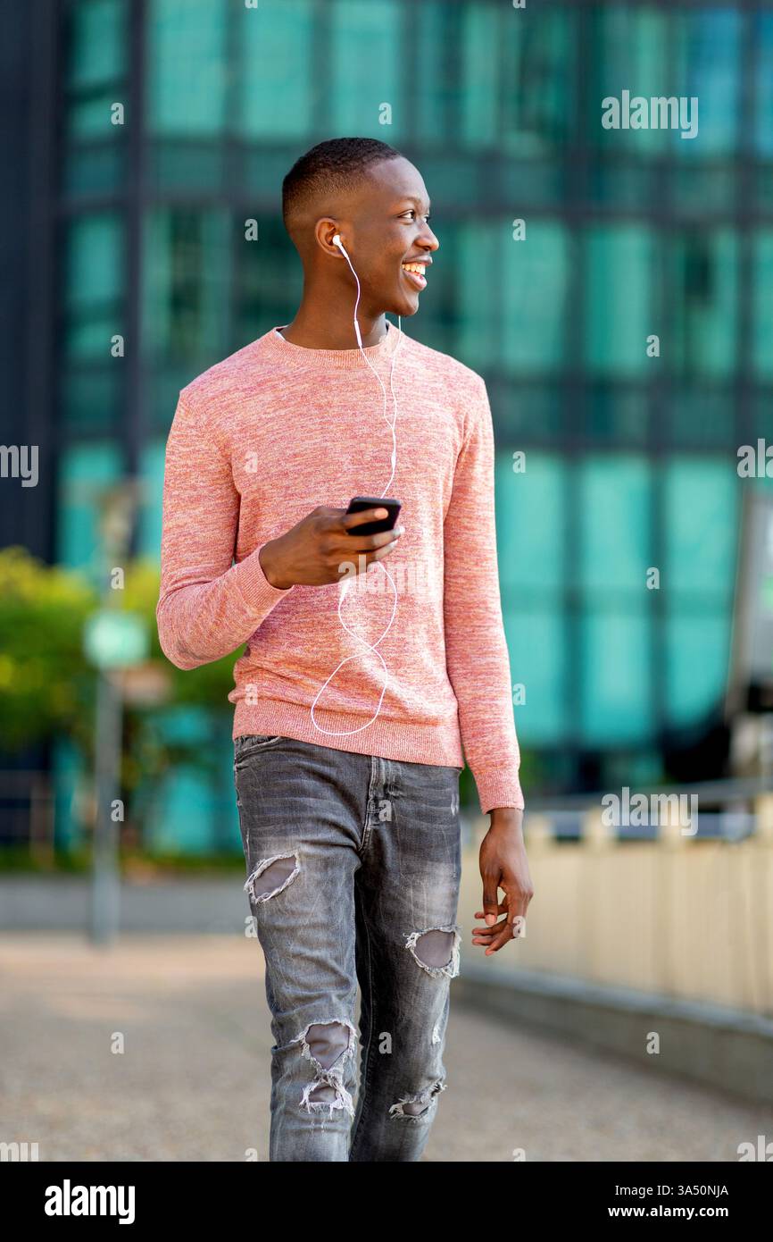 Porträt eines glücklichen jungen Schwarzen Mannes, der durch die Stadt geht und Musik auf seinem Handy mit Ohrhörern hört. Dieses Bild des urbanen Lifestyles zeigt den lässigen Einsatz von Technik und Vergnügen in einer geschäftigen Stadt. Stockfoto