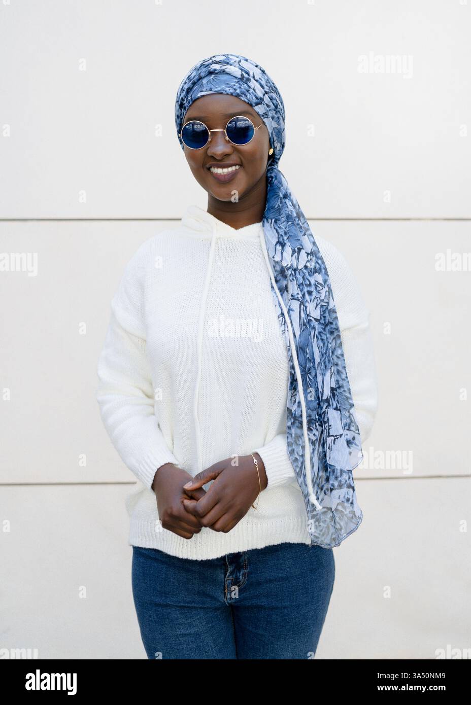 Die lächelnde schwarze Frau mit Kopftuch und Sonnenbrille steht vor einer sauberen weißen Wand. Dieses Mode- und Lifestyle-Porträt betont Stil, Selbstvertrauen und kulturellen Ausdruck. Dank des Kopierbereichs eignet es sich perfekt für die Gestaltung von Marketing- oder Social-Media-Grafiken, die moderne, stilvolle Frauen feiern. Stockfoto