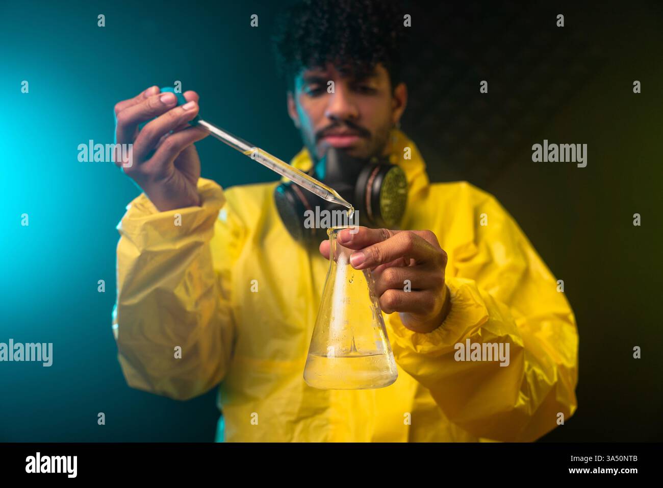 Auffälliges Bild eines männlichen Chemikers aus Spanien in einem Gefahrenanzug und einer Gasmaske, wobei ein Reagenz in einem schwach beleuchteten Labor mit einer Pipette in einen Kolben gegeben wird. Das dunkle Ambiente und die Neonakzente betonen ein wissenschaftliches Hightech-Umfeld. Perfekt für Geschichten über Chemie, Laborsicherheit und Forschung. Stockfoto