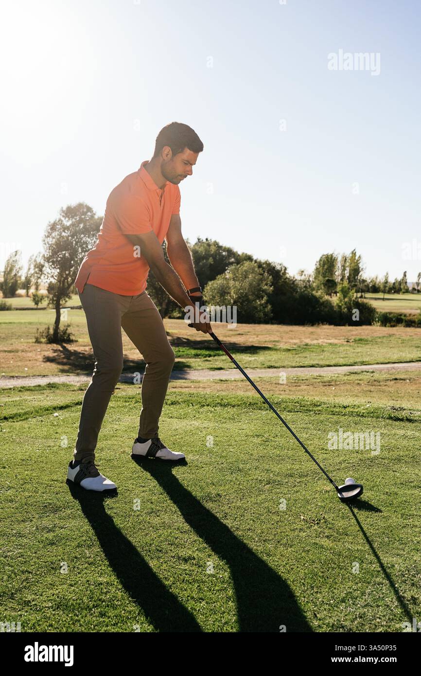 Professioneller männlicher Golfspieler, der sich auf einen grünen Golfplatz vorbereitet, während er auf den Ball hinunterblickt. Das Sommerlicht unterstreicht die grüne Umgebung und die fokussierte Haltung des Athleten. Perfekt für Golf, Sport-Branding und Outdoor-Lifestyle-Kampagnen. Stockfoto