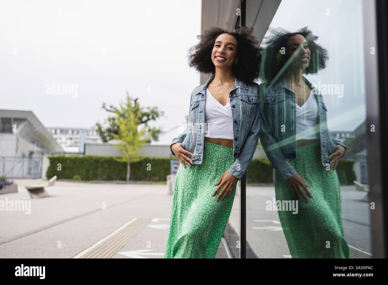 Fröhliche junge afroamerikanische Frau mit lockigem Haar, Jeansjacke und grünem gepunktetem Rock, lächelt in die Kamera, während sie sich an eine Wand lehnt. Ein lebendiges Porträt im Street Style, das Selbstbewusstsein, Mode und urbanes Leben zum Ausdruck bringt. Ideal für Lifestyle, Mode und Accessoires. Stockfoto