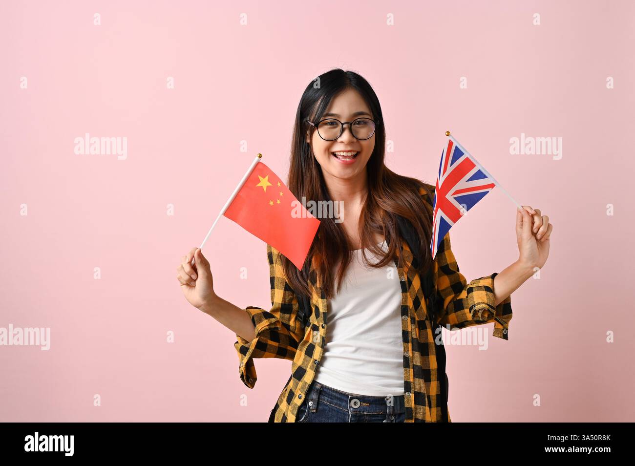 Lächelnde asiatische Studentin in einem Flanellhemd hält England- und China-Flaggen und signalisiert damit ein Auslandsstudium und internationalen Austausch. Dieses Porträt ist isoliert auf einem pinkfarbenen Studiohintergrund und eignet sich für Bildungsmarketing, Hochschulkontakte und Sprachaustauschkampagnen. Das Bild vermittelt Themen wie Jugend, Studium im Ausland und kultureller Austausch. Stockfoto