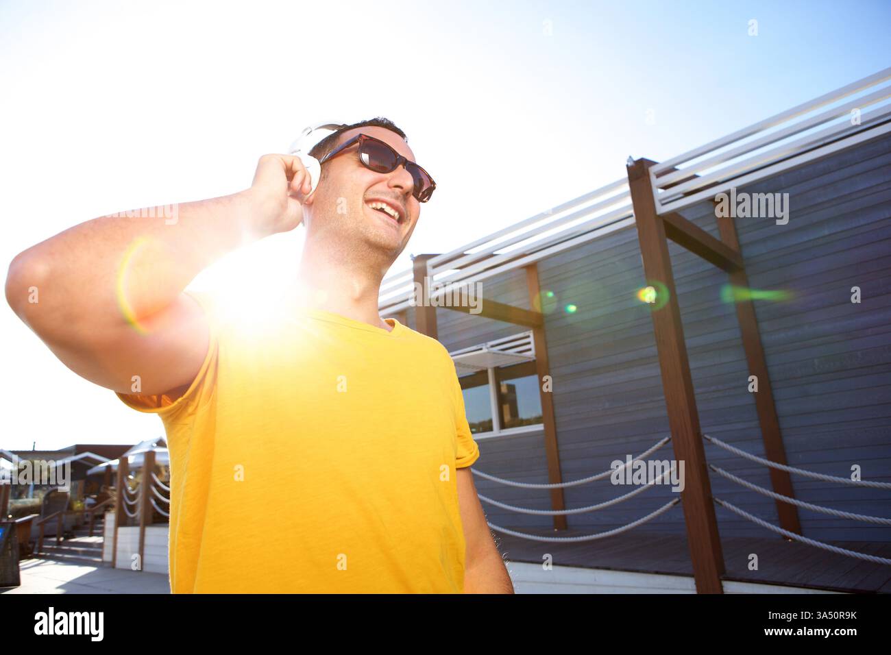 Lächelnder hispanischer Mann mit Handkopfhörern, Musik mit Sonnenbrille hören und am sonnigen Tag im Park wegschauen Stockfoto