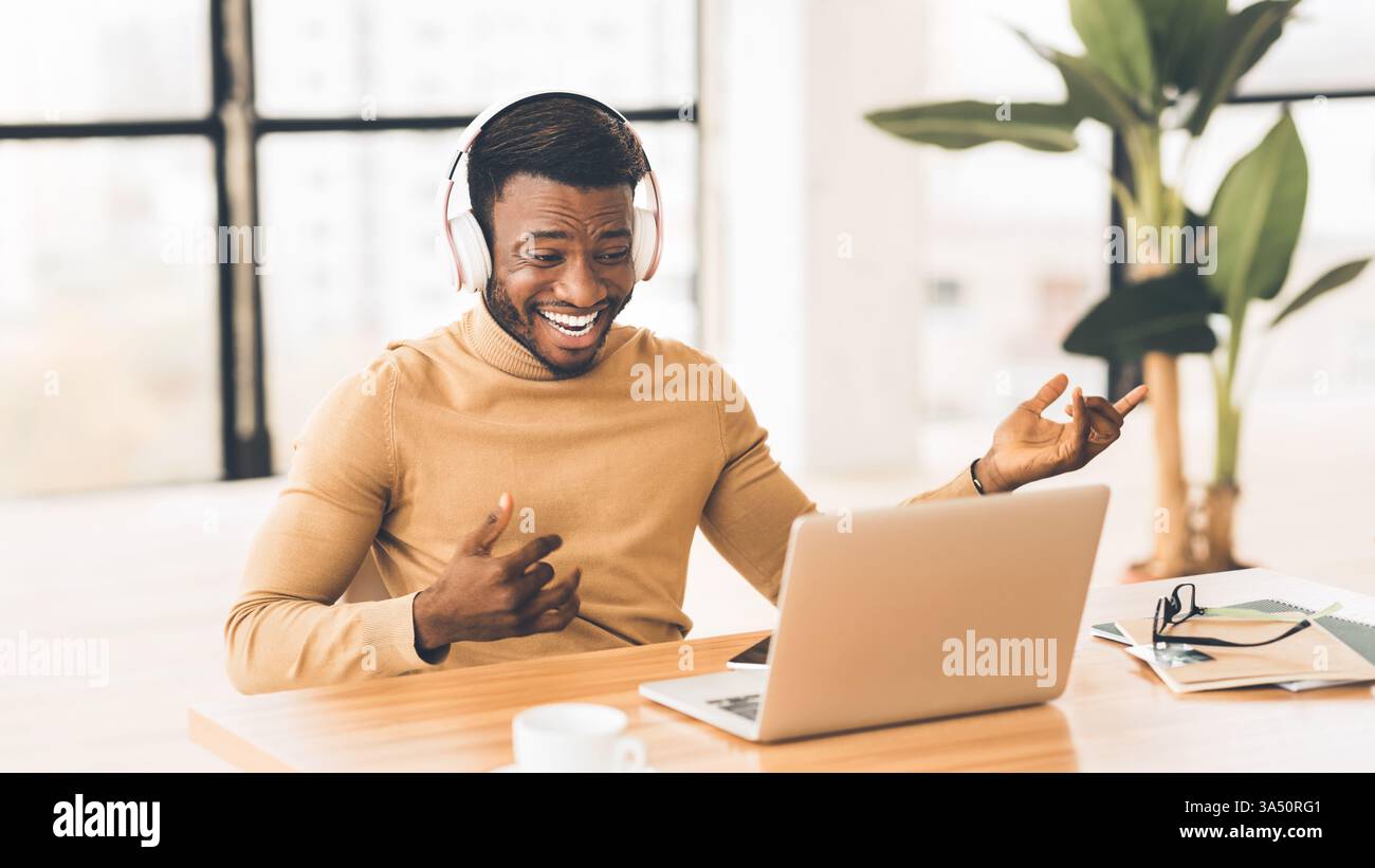 Glücklicher afrikanischer Geschäftsmann hört Musik, während er eine virtuelle Gitarre an seinem Schreibtisch spielt. Panorama-Arbeitsbereich mit Platz für Branding und positiver kreativer Atmosphäre. Ideal für den geschäftlichen Lebensstil, Motivation, Technik- oder Musikkampagnen. Stockfoto