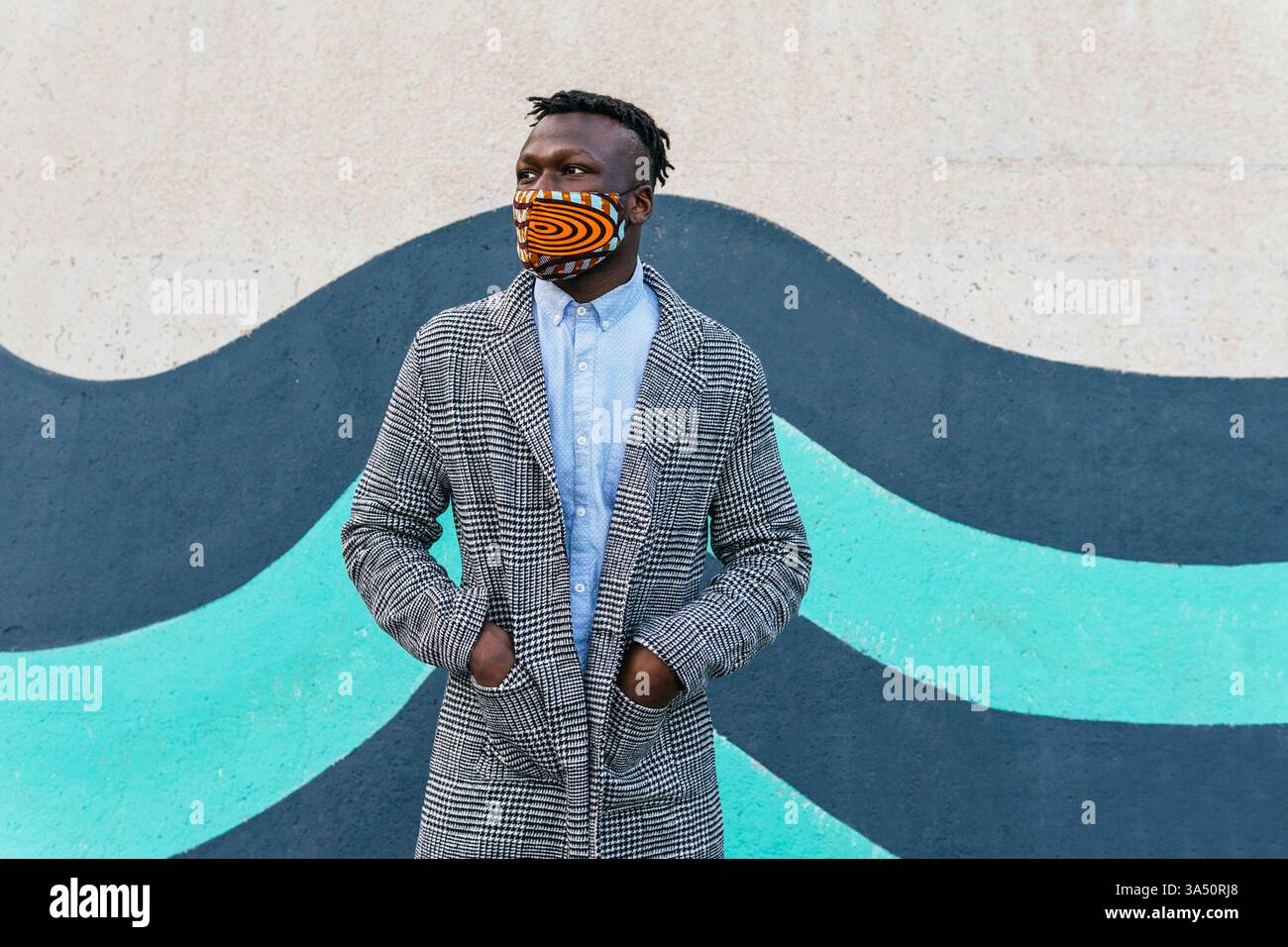 Porträt eines Schwarzen Mannes, der eine Maske trägt und mit Händen in Taschen an einer hellen Wand steht. Dieses Bild spiegelt die COVID-19-Ära und die neue Normalität im öffentlichen Raum wider. Geeignet für den Lebensstil und die Gesundheitskommunikation über Maskengebrauch, Selbstisolierung und vorbeugende Maßnahmen. Stockfoto