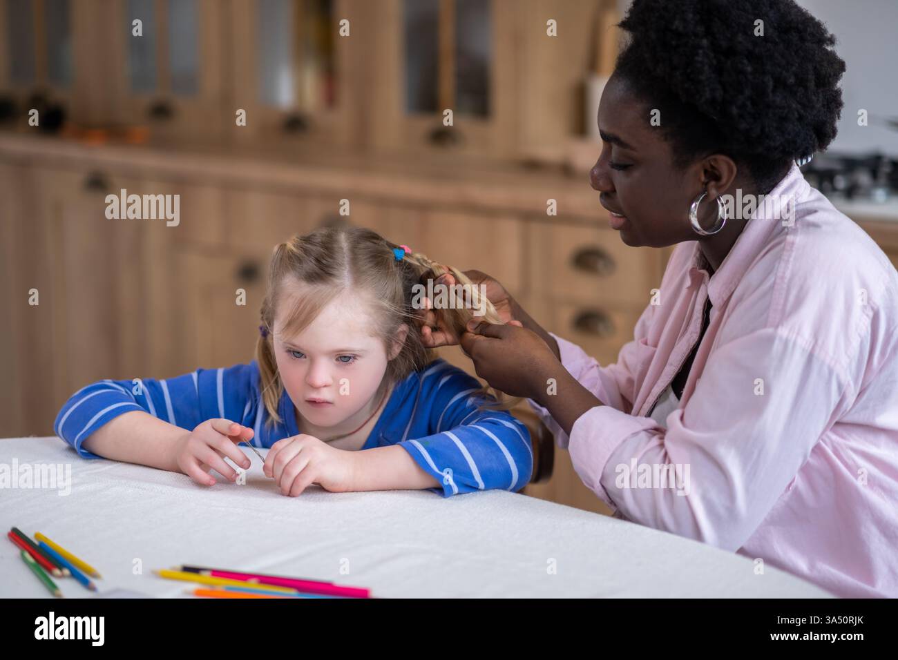 Afroamerikanische Frau, die Haare eines kaukasischen Mädchens mit Down-Syndrom flechtet, das zu Hause am Tisch sitzt Stockfoto