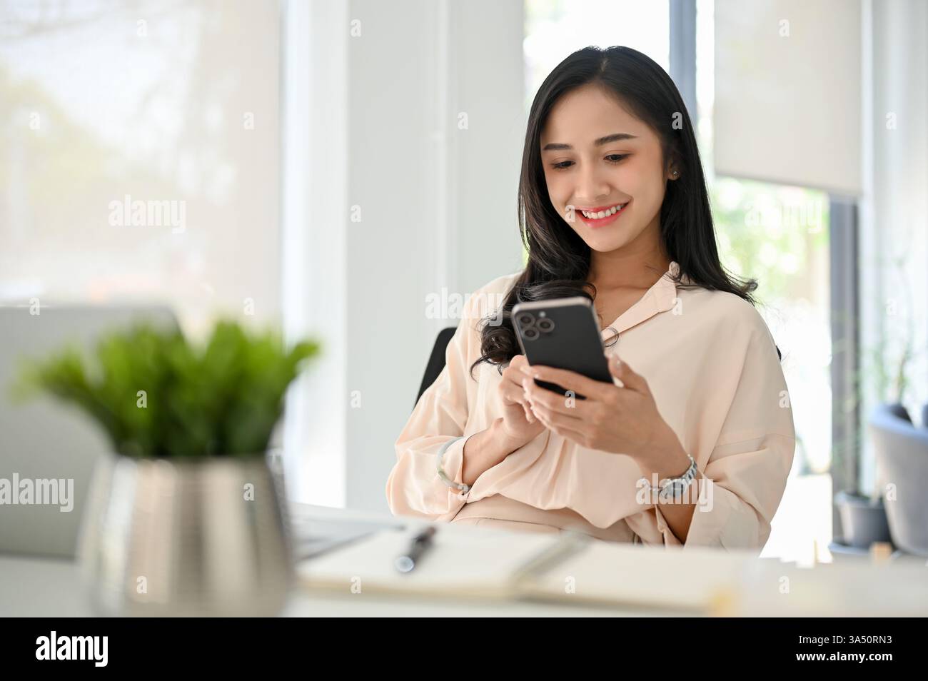 Charmante asiatische Geschäftsfrau mit einem Smartphone an ihrem Schreibtisch in einem modernen Büro. Während der Arbeit chatten und Nachrichten, reflektiert sie moderne digitale Kommunikation in einem professionellen Umfeld. Ideal für geschäftliche, technische und mobile Konnektivität. Stockfoto