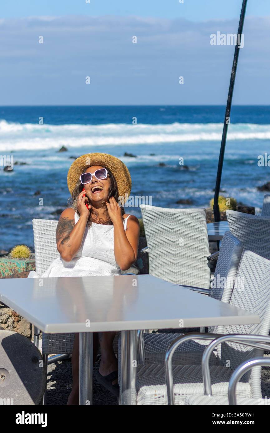 Fröhliche hispanische Lateinfrau lacht, während sie in einem Café am Meer telefoniert. Die Lage am Meer mit blauem Himmel und Blick auf die Küste sorgt für eine entspannte Reise- oder Lifestyle-Stimmung. Ideal für Cafés, Reisen und soziale Medien. Stockfoto
