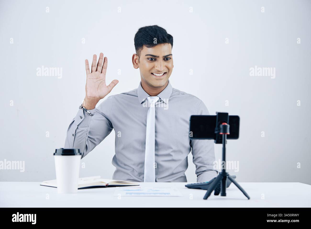 Gut aussehender, lächelnder junger indischer Unternehmer sitzt an einem Schreibtisch mit einer Tasse zum Mitnehmen und einem Kollegen Videoanruf. Die Szene zeigt eine proaktive, moderne Unternehmenskultur und Remote Collaboration in einem modernen Büro. Stockfoto