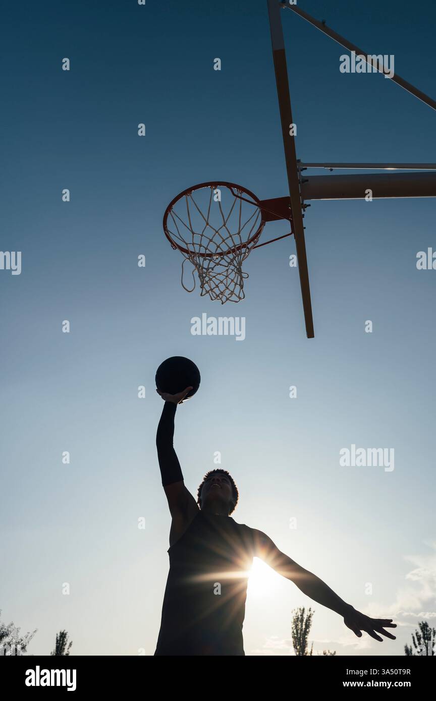 Rückansicht eines energiegeladenen afroamerikanischen Teenager-Basketballspielers, der am Rand hängt, nachdem er auf einem Spielplatz Punkte erzielt hat. Der dynamische Sportschuss vermittelt Energie, Leichtathletik und Jugendwettbewerb. Ideal für Sport, Jugendfitness und Teamkultur. Stockfoto