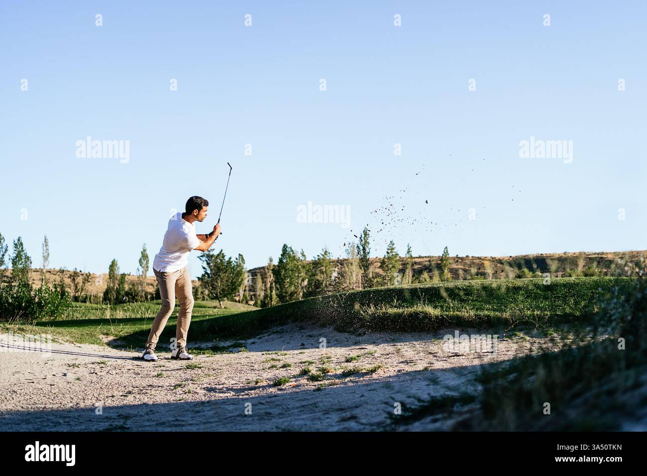 Hispanischer Golfspieler in einem Gefahrenbereich auf einem sonnigen Golfplatz, der wegblickt, während er seinen Schwung vorbereitet. Outdoor-Sportfotografie, die Fokus, Können und Herausforderung hervorhebt. Stockfoto