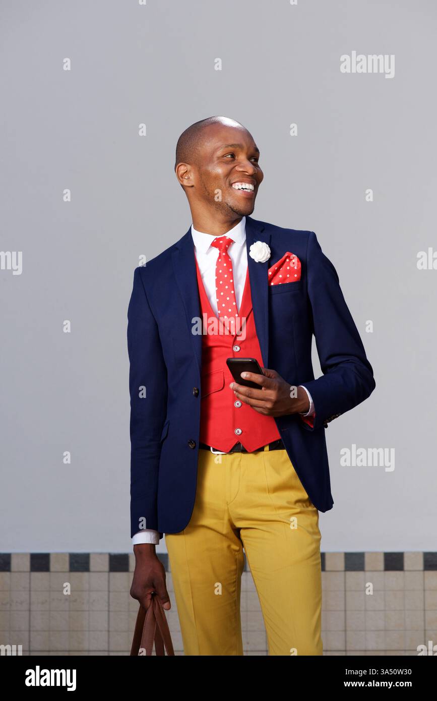 Ein lächelnder afrikanischer Geschäftsmann steht auf der Straße, hält eine Tasche und ein Smartphone. Ein lässiges, aber professionelles Stadtporträt für Unternehmergeist, Networking und Corporate Branding. Ideal für Geschäfts- und Urlaubsreisen. Stockfoto