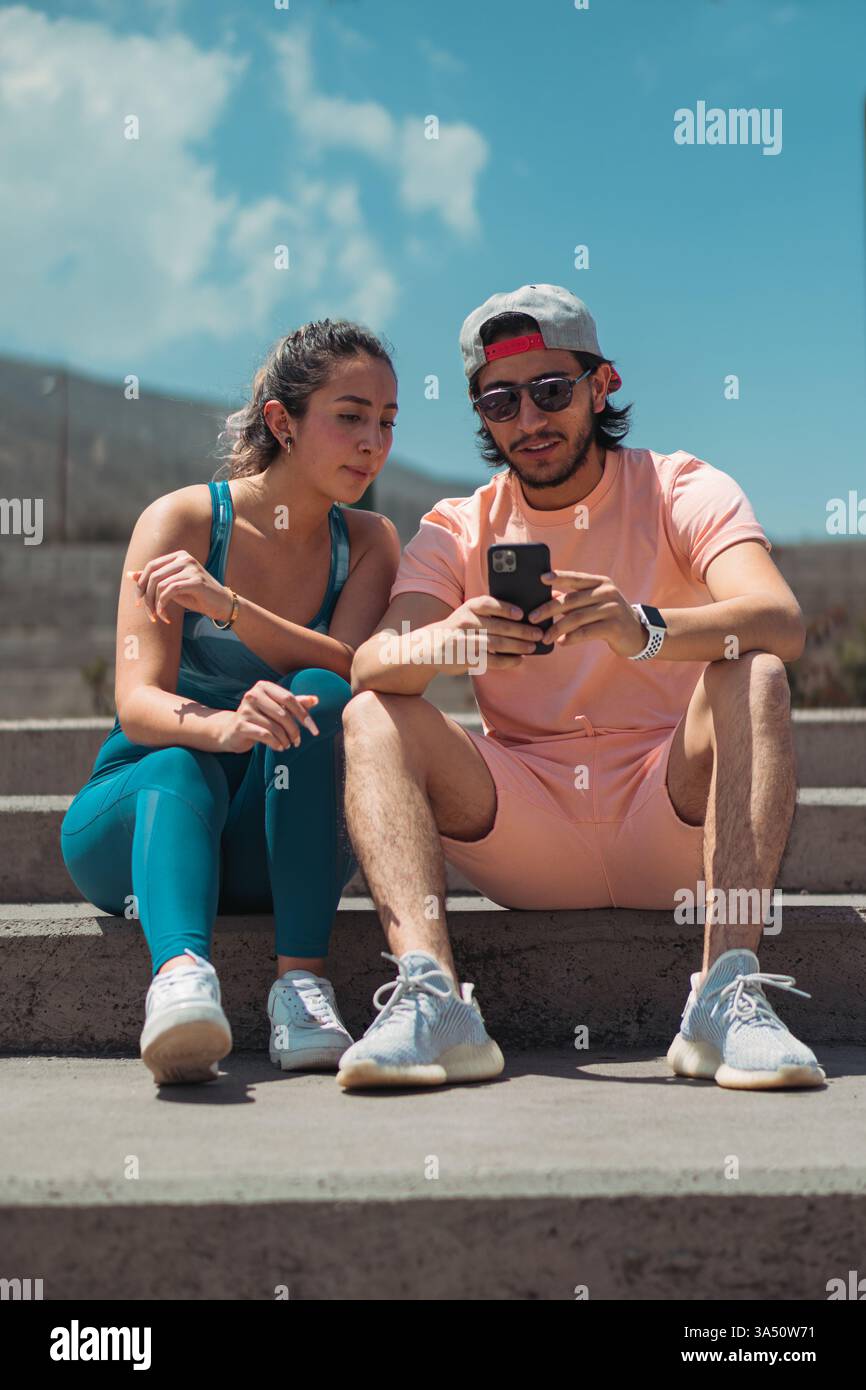 Der hispanische Mann zeigt seiner Freundin mit gemischter Rasse ein Smartphone, während er an einem sonnigen Tag auf einer Parktreppe sitzt. Dies fördert den Lebensstil von Paaren, mobile Technologie und Freizeit im Freien mit warmem, natürlichem Licht. Stockfoto