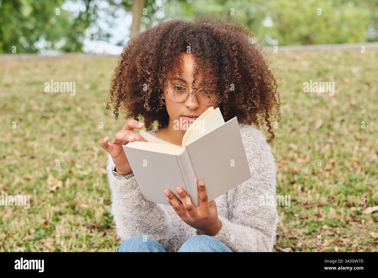 Fokussierte afroamerikanische Frau mit einem Afro liest einen Roman in einem grünen Park, mit einem verschwommenen Hintergrund. Perfekt für Lifestyle-, Lese- und Freizeitkampagnen, die ruhige Momente im Freien hervorheben. Stockfoto
