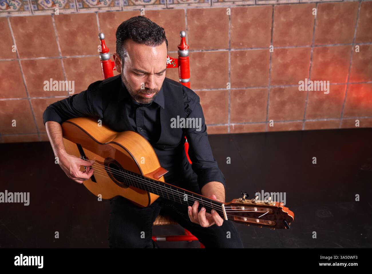 Bärtiger männlicher Musiker in einem schwarzen Rollkragen spielt eine Akustikgitarre drinnen neben einer gekachelten Wand. Ideal für Musik, Performance oder kreative Lifestyle-Kampagnen. Stockfoto