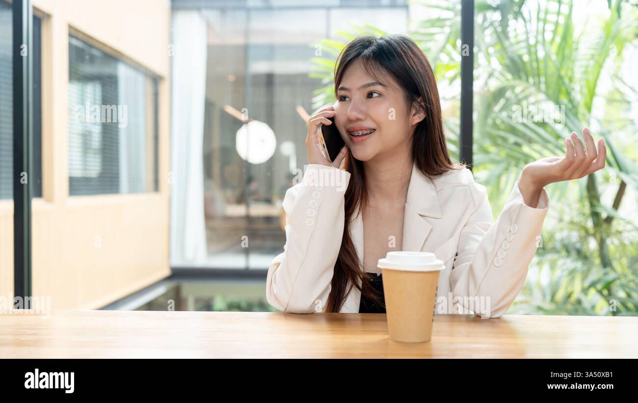 Positive asiatische Geschäftsfrau in einem modernen Loungebereich, die mit einem entspannten Lächeln auf ihrem Smartphone spricht. Die Szene verbindet professionelles Arbeiten mit schnurloser Technologie und lässigem Komfort. Perfekt für die Unternehmenskommunikation, mobile Geschäftsabläufe und moderne Grafik im Büro. Stockfoto