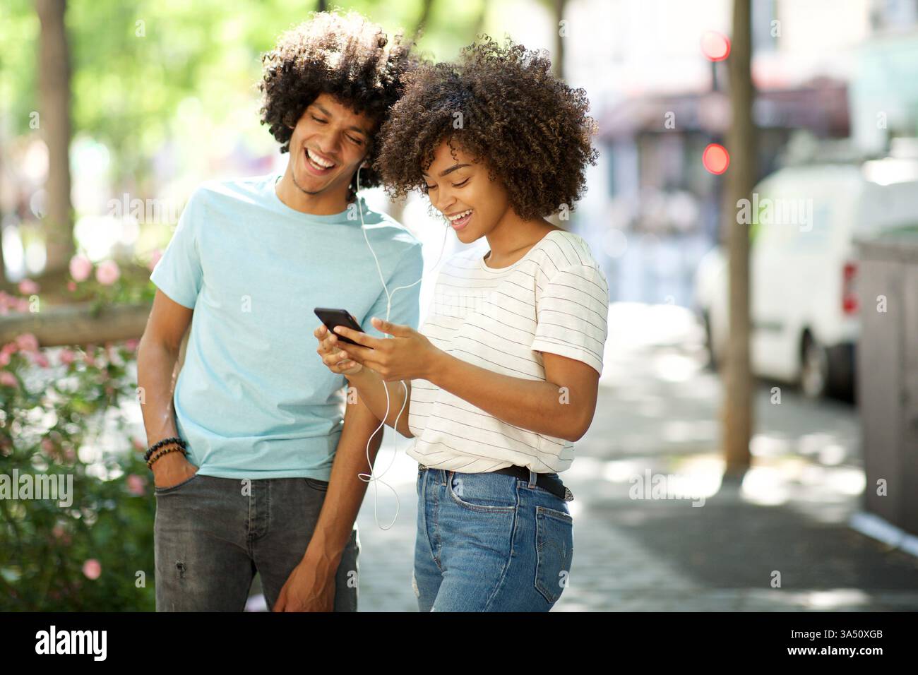 Lächelndes schwarzes Paar, das gemeinsam Musik auf einer Straße genießt. Sie teilen sich Ohrhörer, während sie draußen spazieren gehen und einen lebendigen urbanen Lebensstil zeigen. Stockfoto