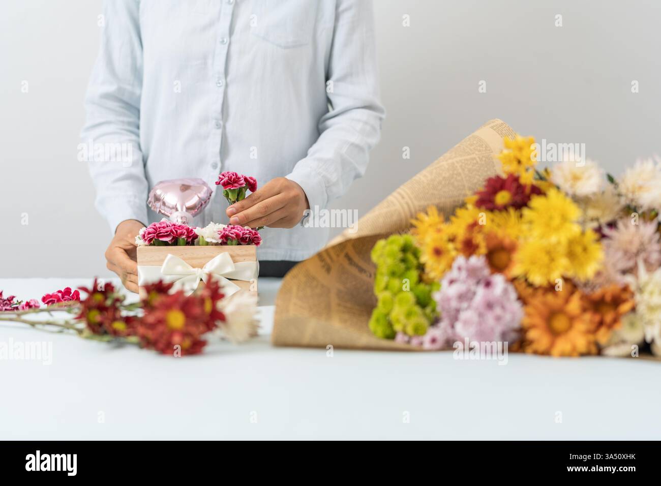 Die hispanische Blumenistin arrangiert rosa und weiße Nelken in einer Holzkiste, die mit einer Schleife in einem Blumengeschäft gebunden ist. Diese fokussierte Komposition unterstreicht das Handwerk und die Präsentation von Geschenken, perfekt für Blumendesign, Hochzeitsdekoration oder Ladenszene. Frische Blüten und warmes Licht sorgen für eine einladende, romantische Stimmung. Stockfoto