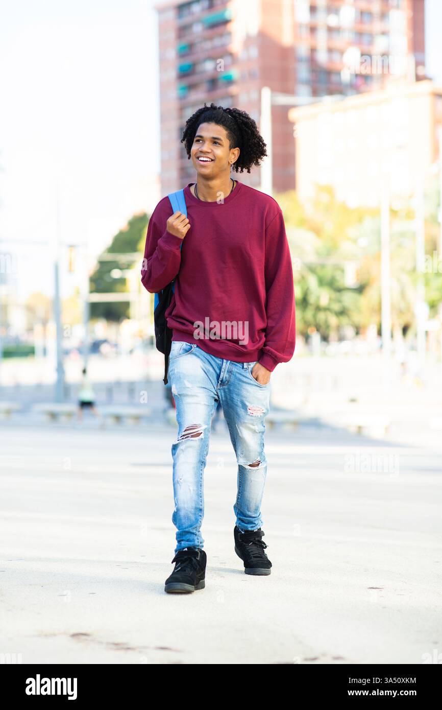 Glücklicher afroamerikanischer Mann, der draußen in der Stadt mit einem Rucksack spaziert. Lässige urbane Lifestyle-Szene, die Bewegung, Stil und tägliche Entdeckungen hervorhebt. Geeignet für Reisen, Pendelfahrten und Jugendstilkampagnen. Stockfoto