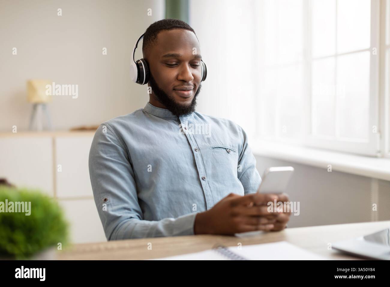 Entspannter Schwarzer Business Professional mit Smartphone und Kopfhörern an einem modernen Schreibtisch. Die Szene zeichnet eine ruhige, produktive Stimmung auf, die sich für die Themen Technologie, Arbeitsleben und Remote Collaboration eignet. Ideal für Unternehmenskommunikation, Apps und Kampagnen mit Musik- oder Podcast-Bezug. Stockfoto