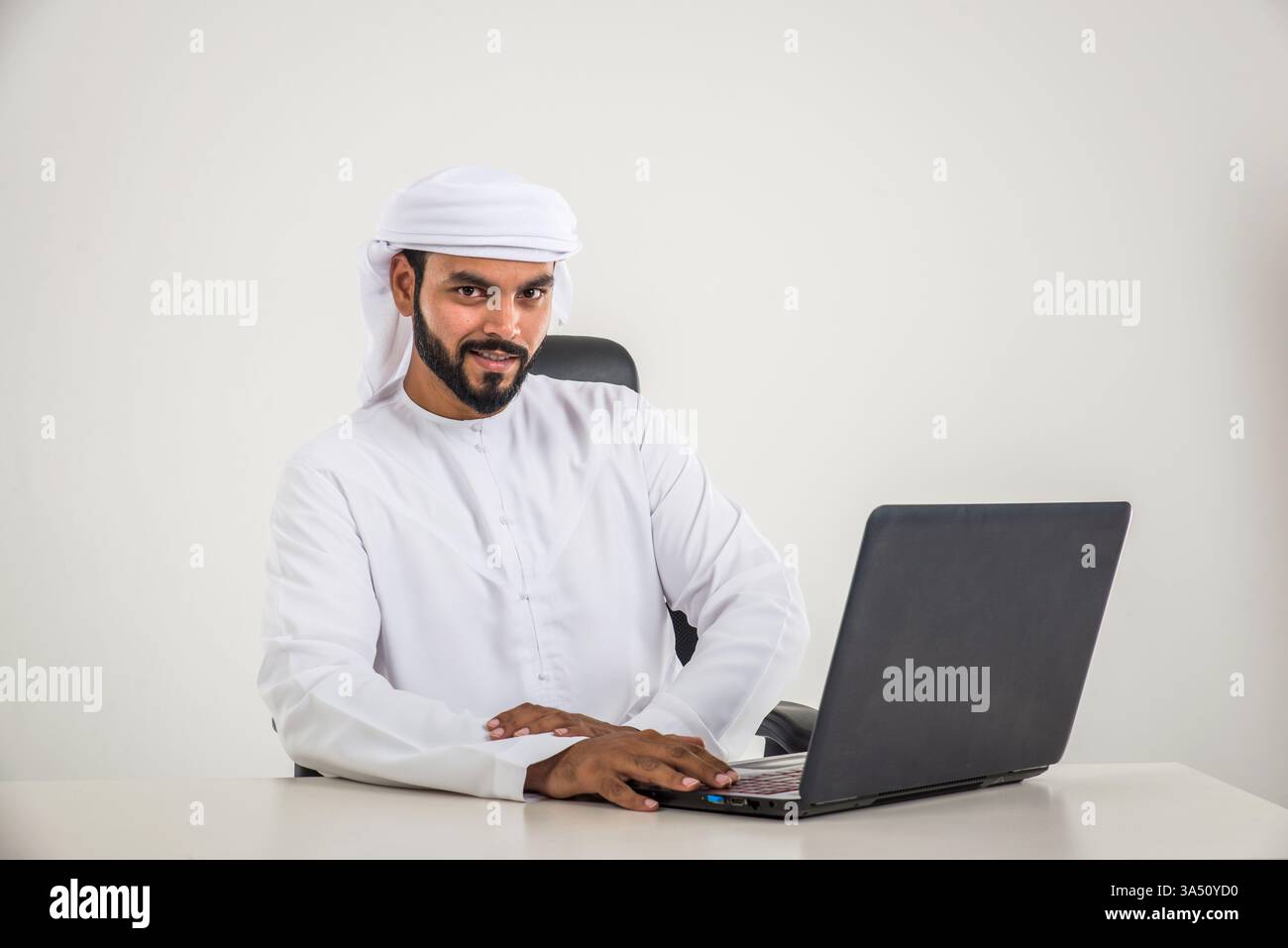 Ein lächelnder Mann mit traditioneller Kandura posiert in einem Fotostudio. Dieses Lifestyle-Porträt spiegelt die zeitgenössische Kultur und professionelle Präsentation der Emirate wider. Geeignet für Lifestyle-, Geschäfts- und Familien- oder Kulturkampagnen im Nahen Osten. Stockfoto