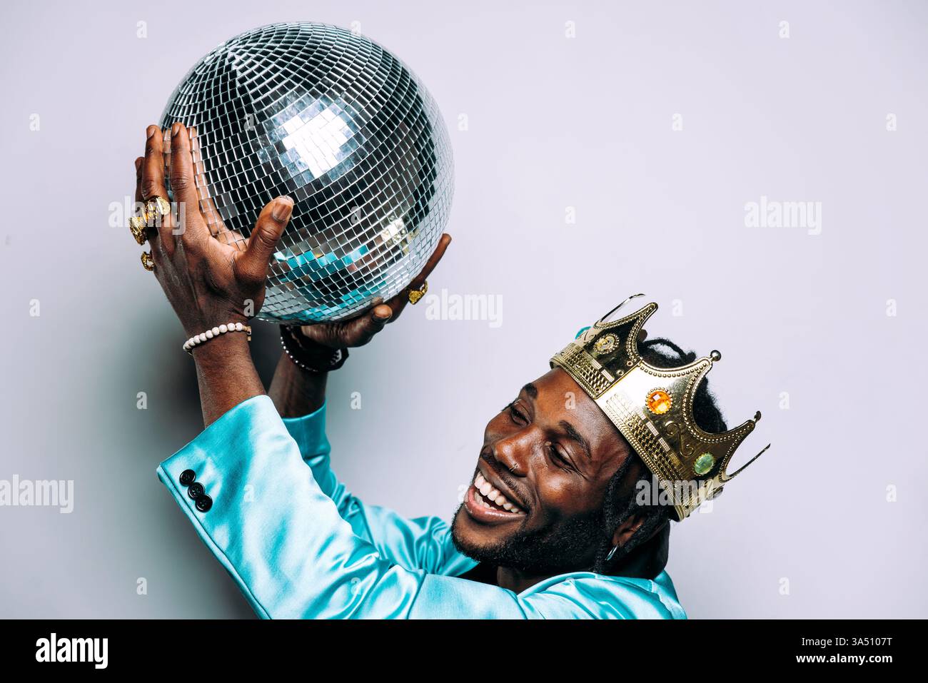 Ein lächelnder Afroamerikaner, der eine Krone trägt, hält eine Disco-Spiegelkugel vor einem grauen Studio-Hintergrund. Das verspielte Porträt vermittelt Feier und kreativen Stil. Stockfoto