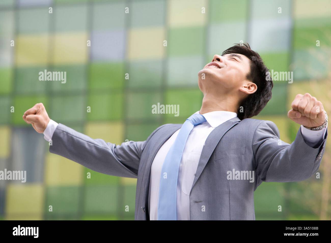 Fröhlicher chinesischer Geschäftsmann im Freien, der mit gezogenen Fäusten vor einem Bürogebäude aufblickt. Ein lebendiges Bild für Leistung, Motivation und Erfolg. Stockfoto