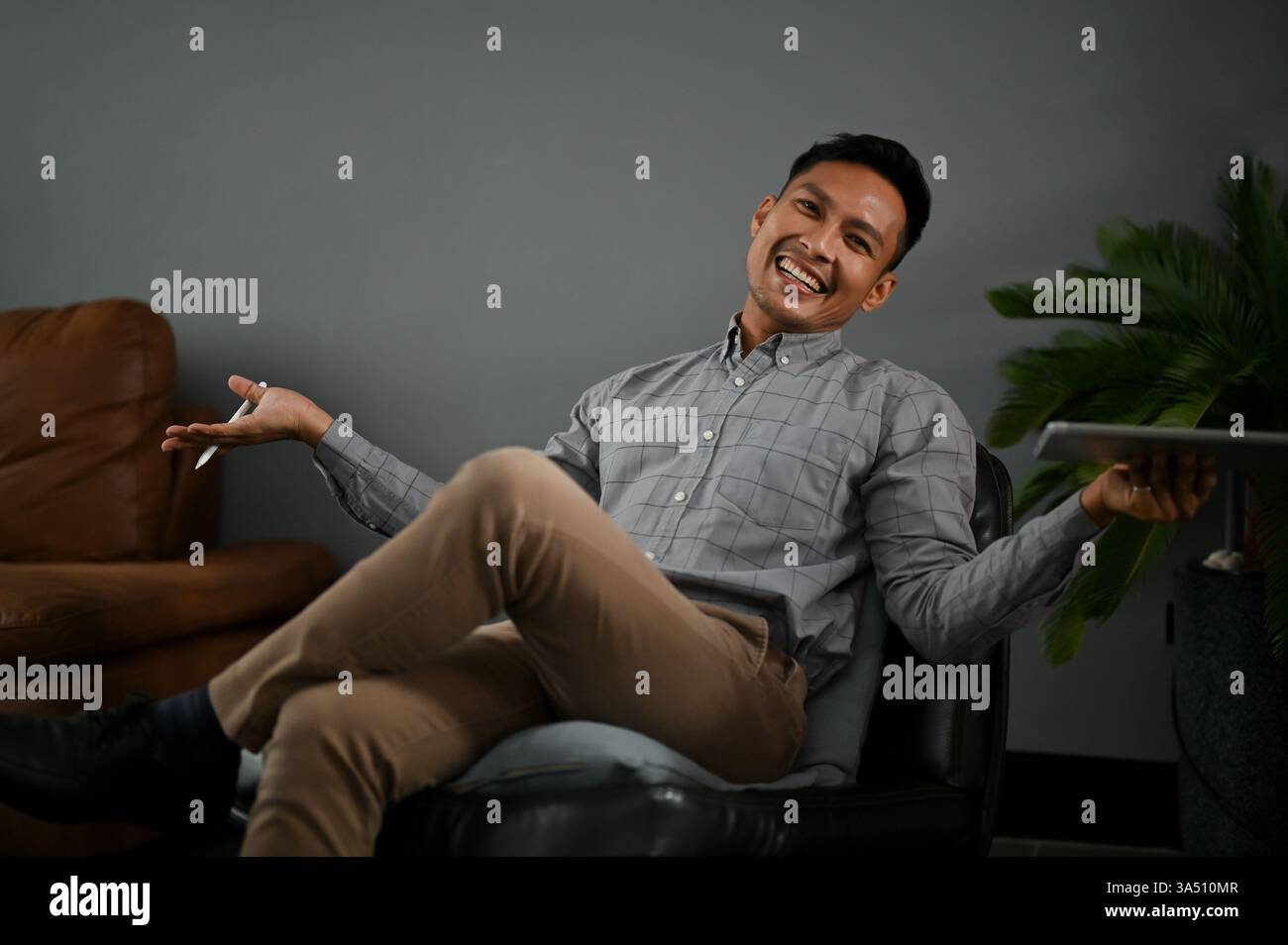 Fröhlicher tausendjähriger asiatischer Mann in lässiger Kleidung in einem modernen Wohnzimmer auf einem schwarzen Ledersessel sitzend, die Handflächen offen in einer sorglosen Pose. Ideal für Lifestyle, Unternehmergeist und Arbeitsplatzkonzepte mit entspannter Atmosphäre. Ideal für Artikel über Vertrauen, Führung und moderne Büros. Stockfoto