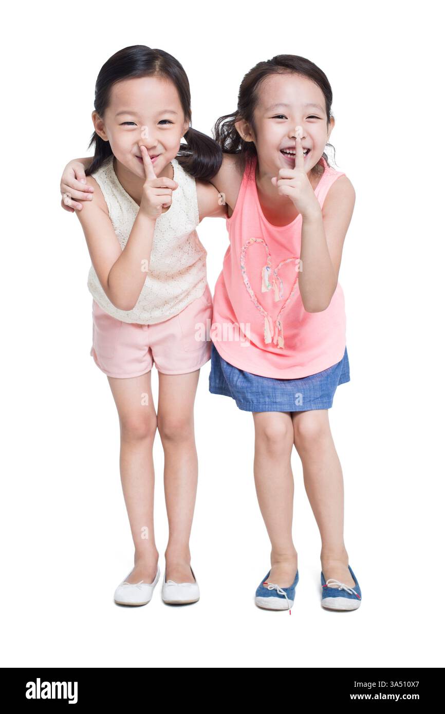 Glückliche chinesische Mädchen mit Fingern auf den Lippen teilen sich einen verspielten Moment drinnen. Vielseitig einsetzbar für Freundschaft, Kindheit und unbeschwerte Lifestyle-Themen. Stockfoto