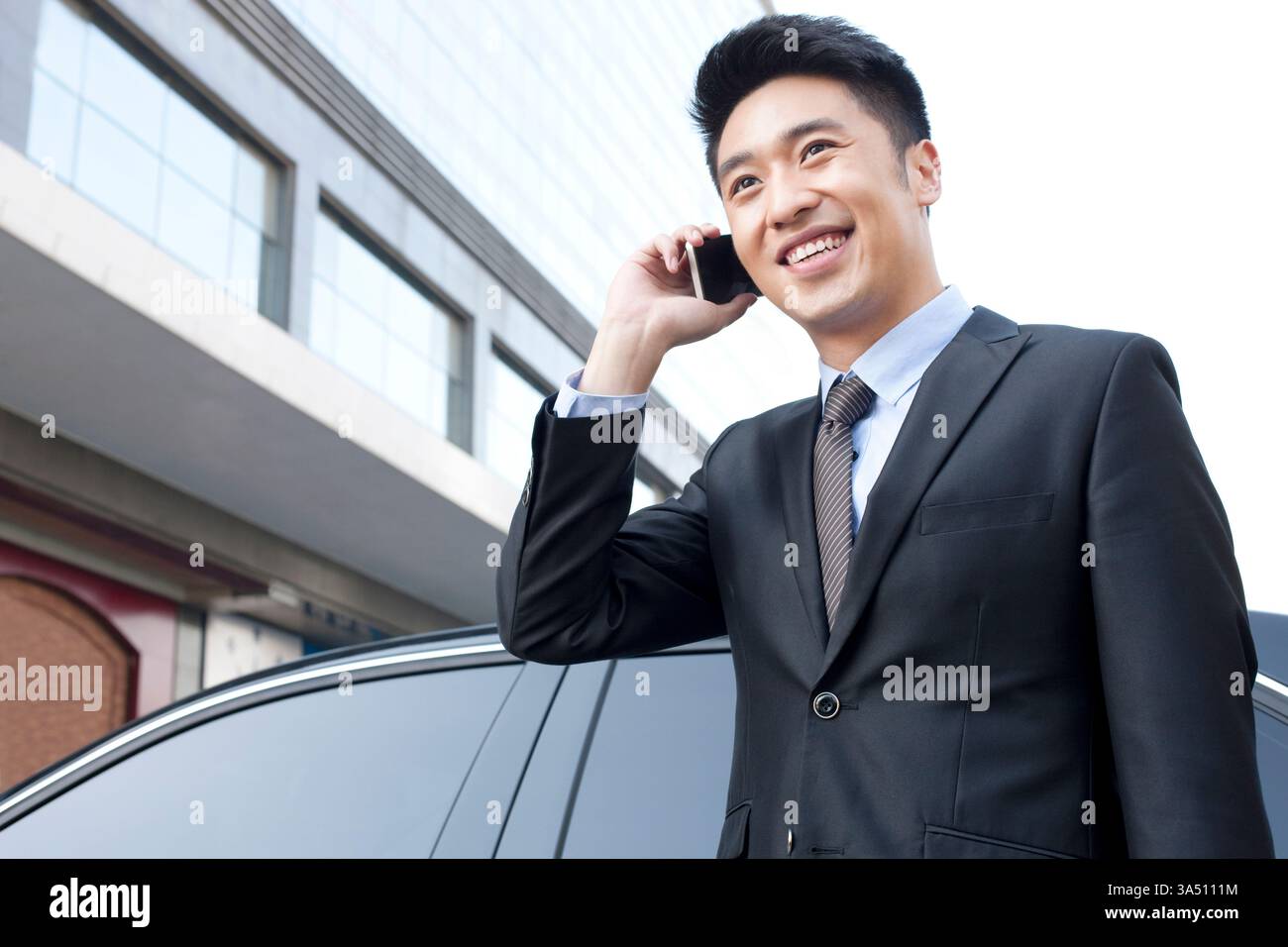 Fröhlicher chinesischer Geschäftsmann am Telefon neben einem Auto in einem belebten Stadtteil in Peking, China. Dieser geschäftliche Moment passt zum Stadtleben, zu Business und Startup-Visuals in China. Stockfoto