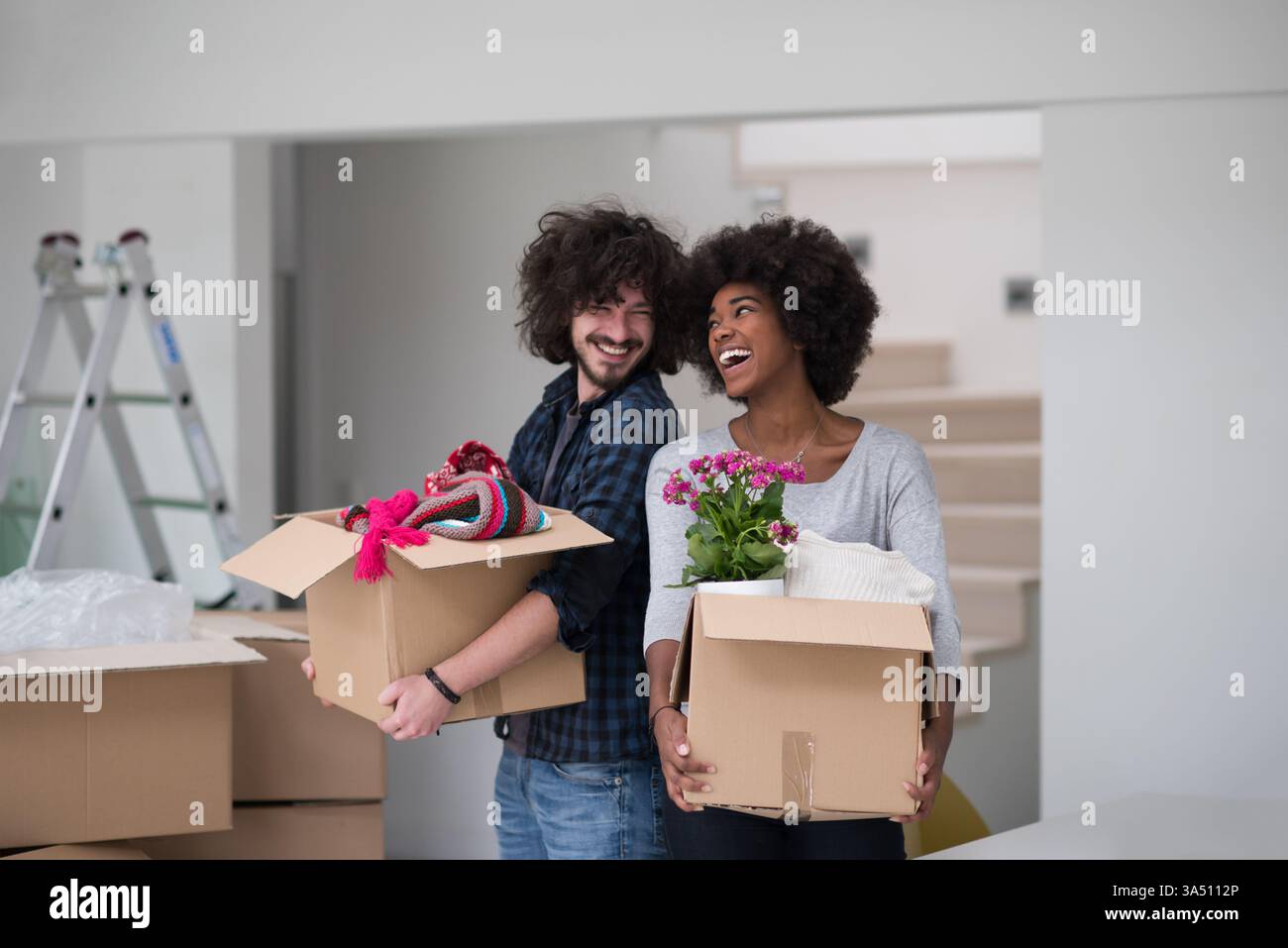 Fröhliches, multiethnisches Paar packt Kartons aus, während sie in ein neues Zuhause ziehen. Eine warme Szene, die sich perfekt für Geschichten über Umsiedlung, Umzugstag und den Beginn des gemeinsamen Lebens in einem stilvollen Raum eignet. Stockfoto