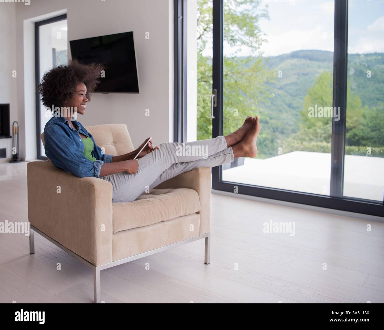 Junge afroamerikanische Frau zu Hause, die sich in einem luxuriösen Wohnzimmer entspannt und ein Tablet liest und im Internet surft. Dieses moderne Lifestyle-Bild eignet sich für Fernarbeit, digitale Unterhaltung und private Komfortkampagnen. Perfekt für Funktionen zum Online-Lernen, zur Freizeit und zur gehobenen Innenausstattung. Stockfoto