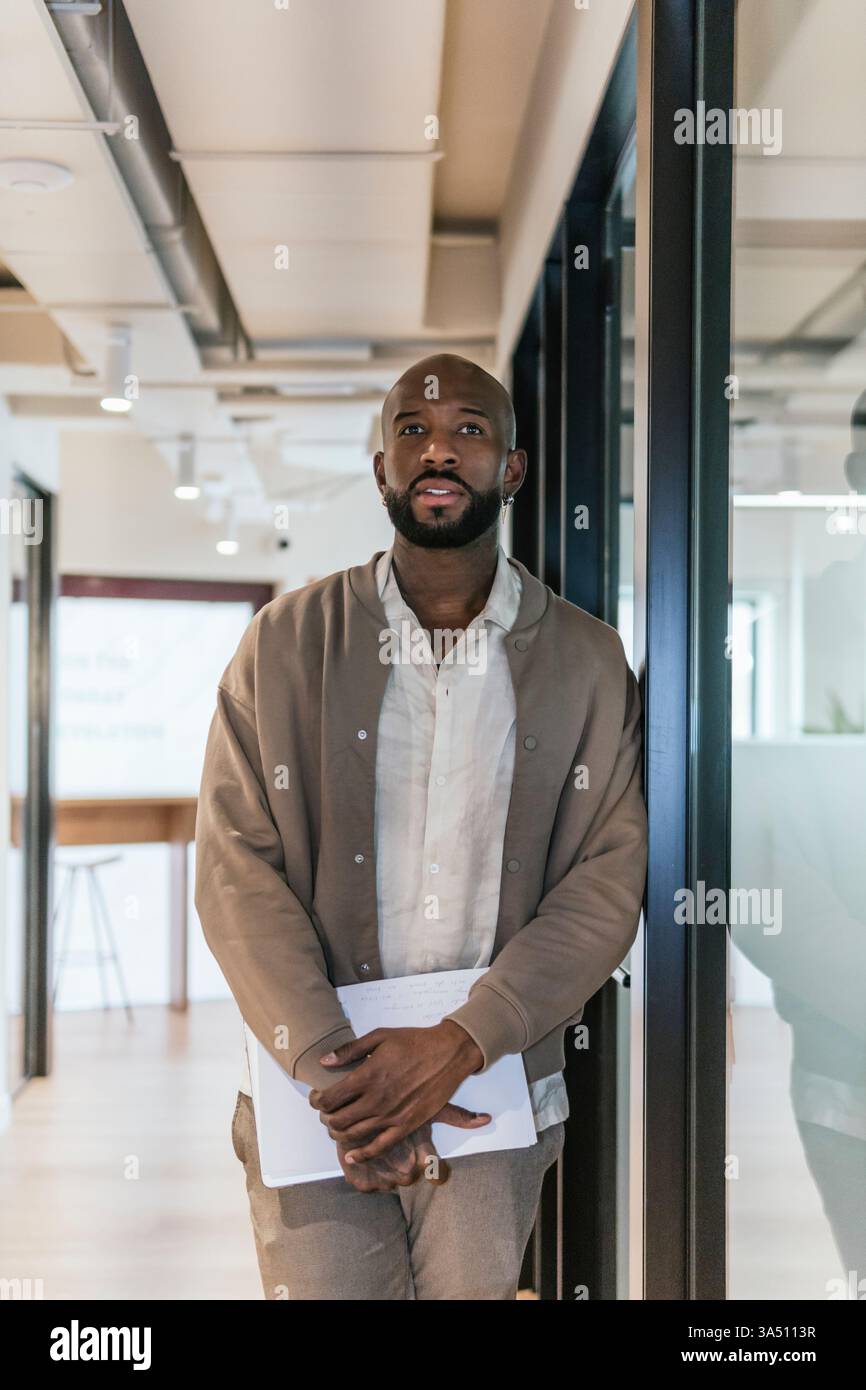 Ein gut gekleideter Afroamerikaner steht kontemplativ in einem modernen Coworking-Büro und hält Dokumente. Die stilvolle Büroszene vermittelt Professionalität und Konzentration in einer modernen Arbeitsumgebung. Ideal für Business-, Start-up- und Büroinhalte mit einem selbstbewussten Profi. Stockfoto
