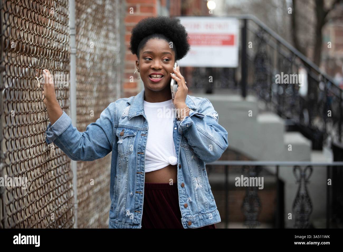 Junge afroamerikanische Frau in einer Jeansjacke, die einen Zaun in einer städtischen Umgebung berührt. Ein lässiges, modernes Lifestyle-Image, das sich für Mode, Street-Style und Alltagsbewusstsein eignet. Stockfoto