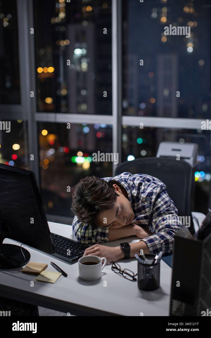 Ein müder chinesischer Mann in einem karierten Hemd schläft ein, während er nachts spät im Büro arbeitet. Dieses Bild vermittelt Überstunden, IT- oder Bürostress und nächtliche Arbeitsrealitäten – ideal für Technologie- und Geschäftsabdeckungen. Geeignet für Geschichten über lange Stunden und Burnout am Arbeitsplatz. Stockfoto