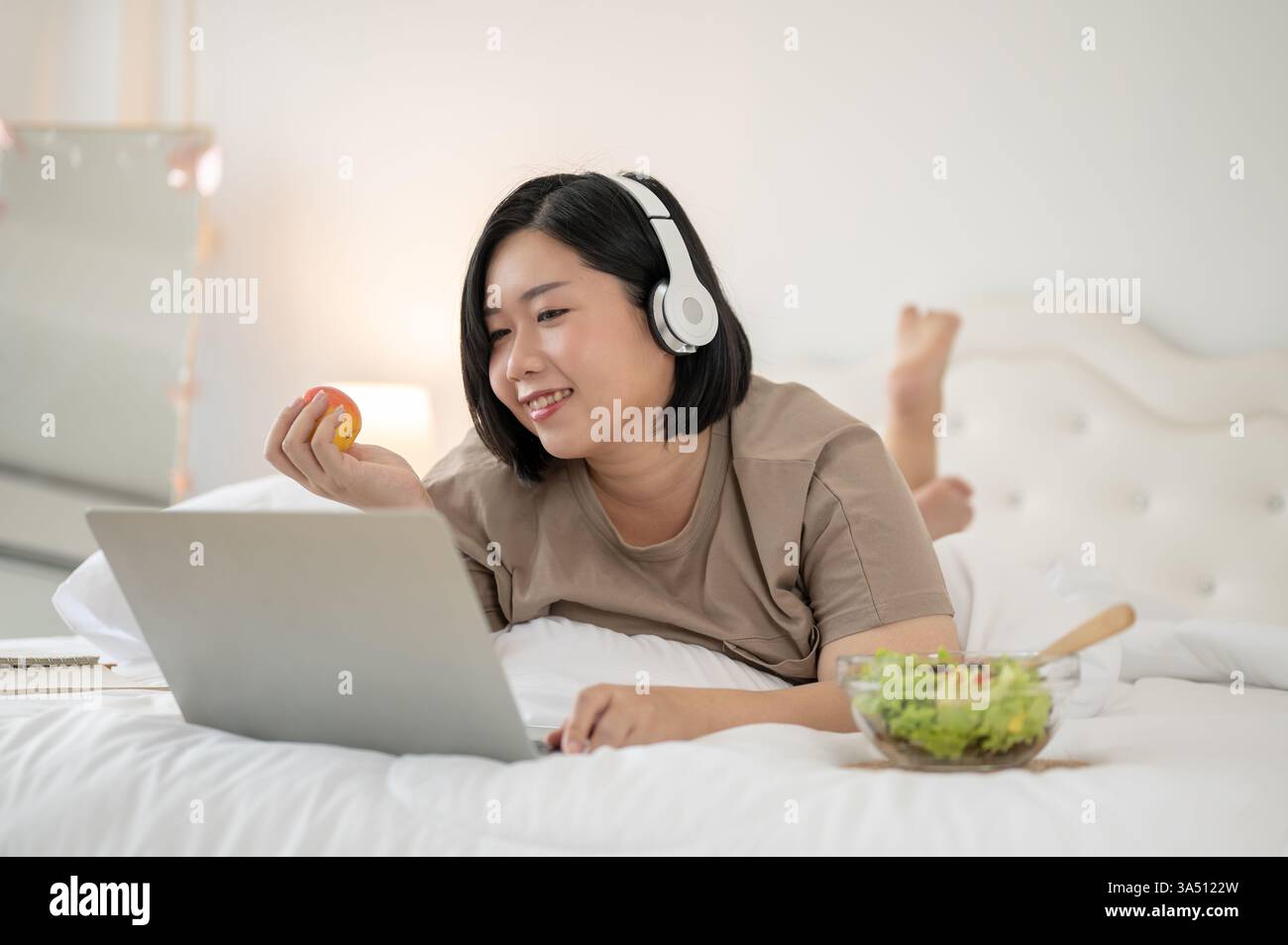 Glückliche asiatische Frau in Übergröße, die sich im Bett mit Kopfhörern und einem Laptop entspannt. Sie schaut sich einen Film an, surft online und plaudert aus einem gemütlichen Schlafzimmer. Perfekt für Lifestyle, Fernarbeit und Selbstpflege. Stockfoto