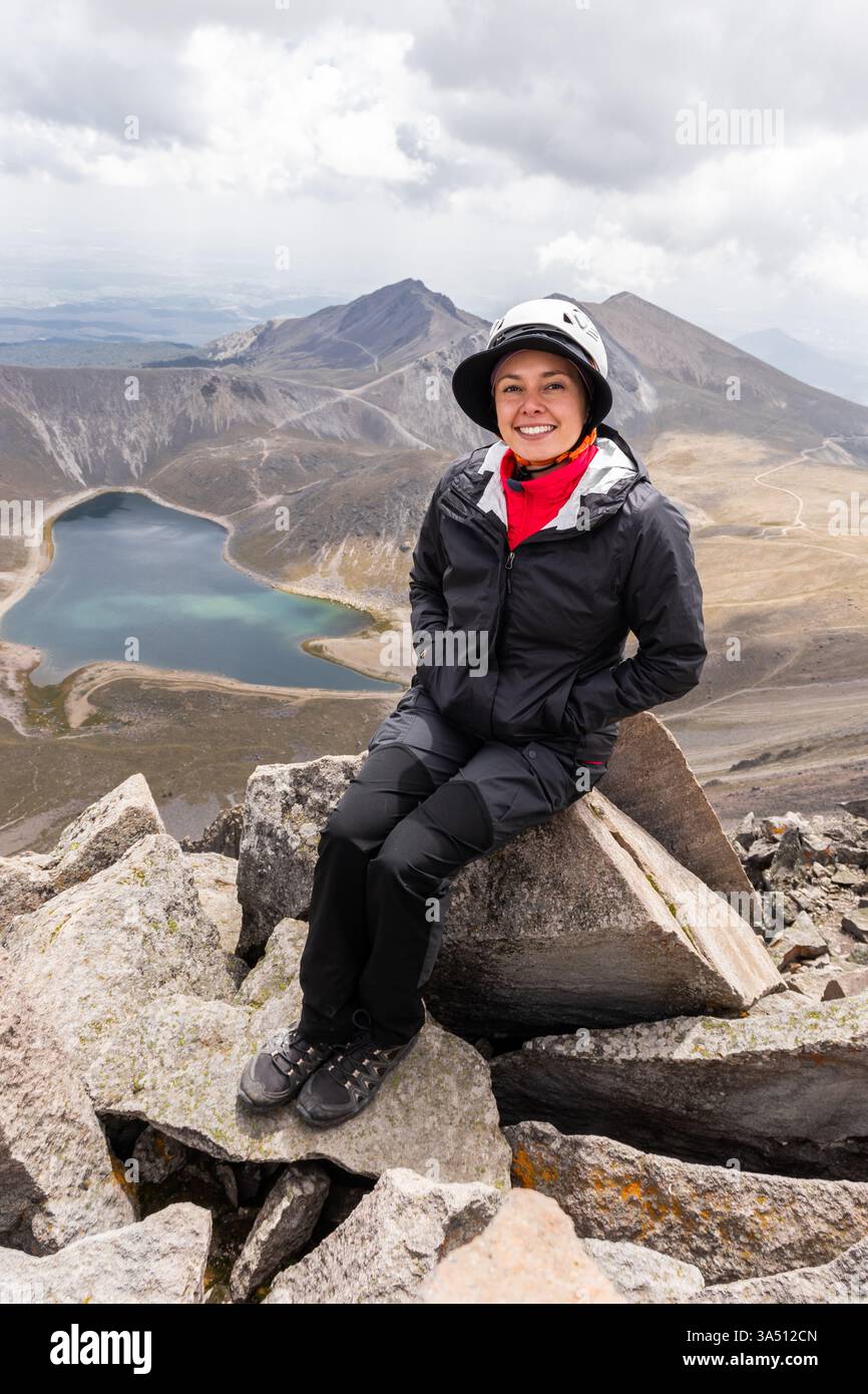 Ein fröhlicher Hispanic Wanderer sitzt auf einem Felsvorsprung mit Blick auf einen vulkanischen Kratersee in Mexiko. Diese Outdoor-Szene zeigt Abenteuer und Natur mit einem dramatischen Blick auf den Krater, den See und das zerklüftete Gelände, perfekt für Reisen, Wandern und Erkundungsthemen. Stockfoto