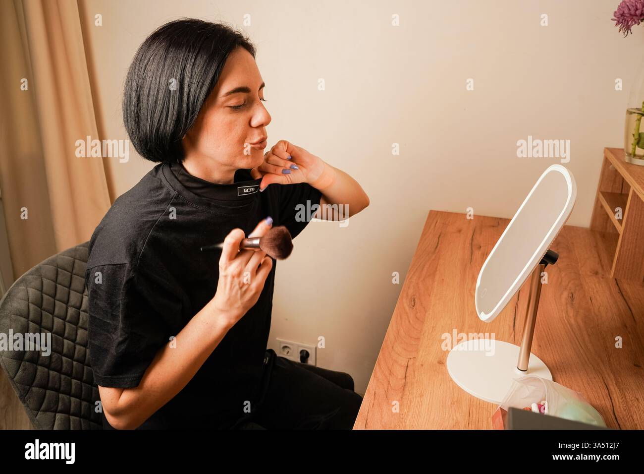 Ihre zarten Züge spiegeln sich im Tischspiegel wider. Ihr dunkles Haar rundet ihren Look ab. Die Frau konzentriert sich auf ihr Make-up und benutzt einen weichen Pinsel Stockfoto
