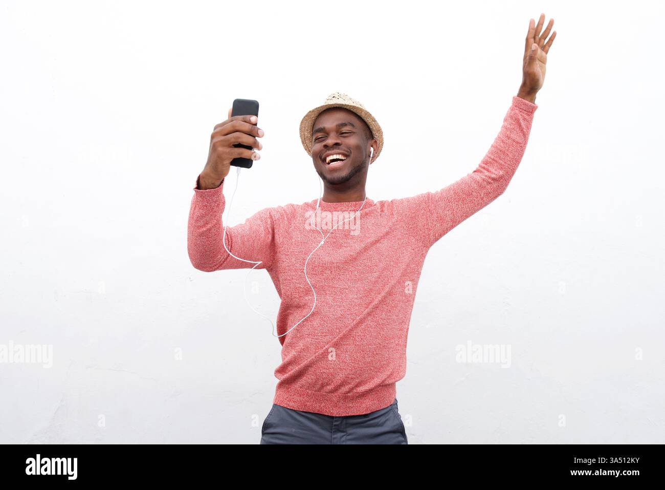 Porträt eines glücklichen afroamerikanischen Mannes mit erhobenem Arm, während er Musik auf seinem Telefon mit Ohrhörern hört. Diese energiegeladene Lifestyle-Aufnahme betont Freude, Bewegung und moderne Technik. Ideal für Unterhaltung, Musik und Jugendkampagnen. Stockfoto