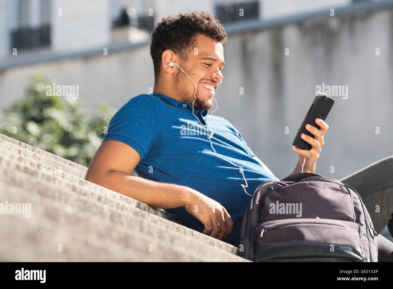 Nahaufnahme des Porträts eines glücklichen jungen afroamerikanischen Mannes, der auf Stufen in einer urbanen Umgebung sitzt und Musik auf einem Smartphone mit Ohrhörern hört. Diese legere Lifestyle-Aufnahme passt zu Streetwear, Stadtleben und mobilen Technologien. Eine Tasche sorgt für alltäglichen Kontext. Stockfoto