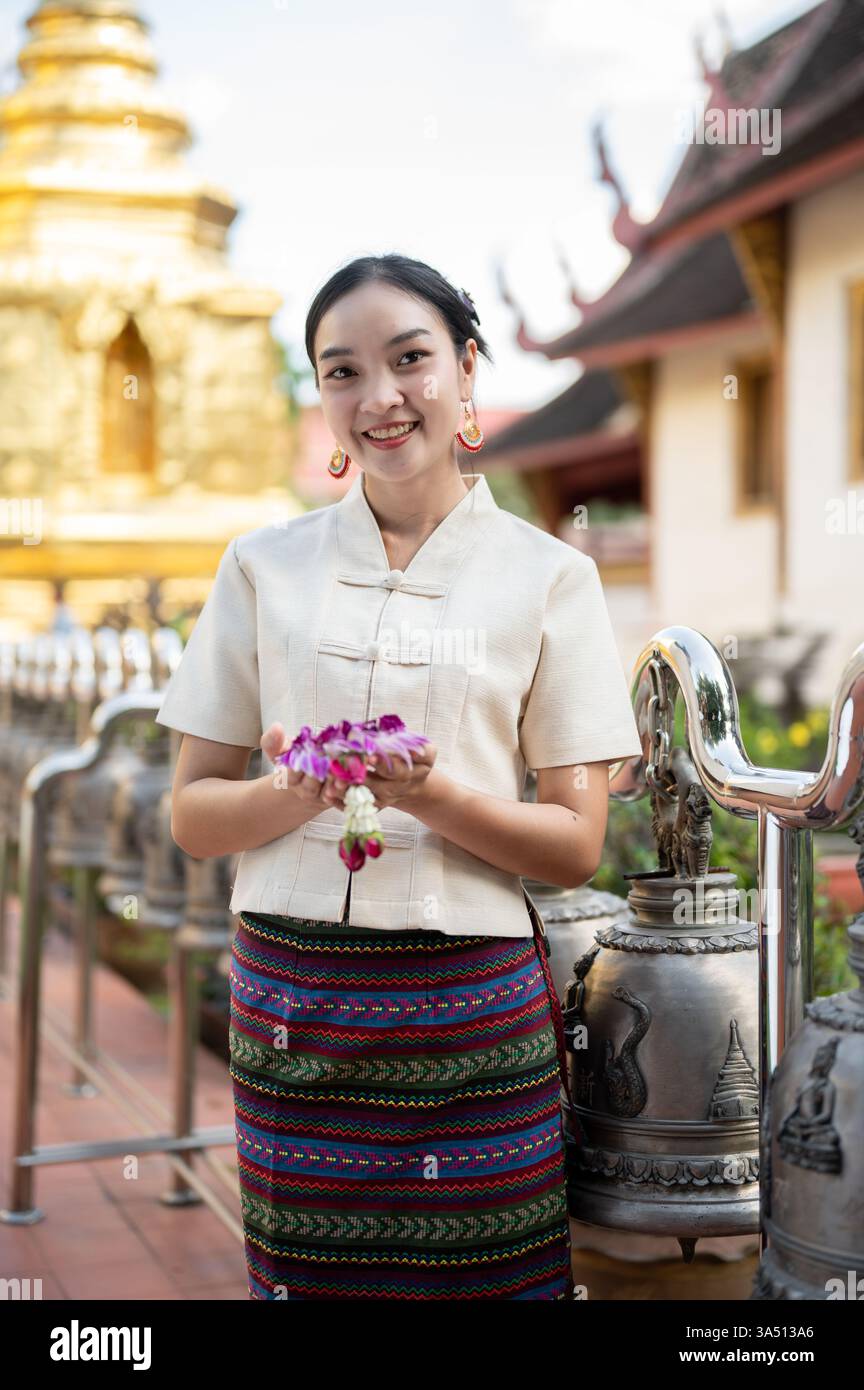 Eine lächelnde Thai-Frau in traditionellem nordischem Kleid hält eine Girlande in einem Tempel. Das Bild hebt das kulturelle Erbe, das Tempelambiente und die buddhistische Tradition hervor. Ideal für Reisen, Kultur und spirituelle Lifestyle-Kampagnen. Stockfoto