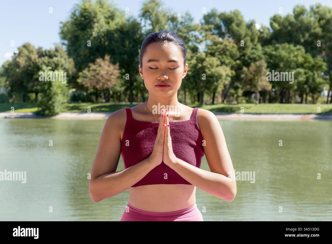 Asiatische junge Frau in Sportbekleidung spielt eine Namaste an einem ruhigen See, umgeben von grünen Bäumen. Dieses Wellness-Image ist ideal für Yoga, Achtsamkeit und Outdoor-Fitness-Kampagnen. Stockfoto