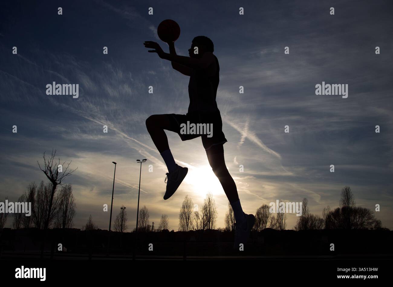 Ganzkörper-Silhouette eines gesichtslosen männlichen Athleten in Sportswear, der abends mit einem Basketball auf einem Spielplatz springt. Die stimmungsvolle Beleuchtung betont sportliche Action und Outdoor-Sport-Energie. Dieses Image eignet sich gut für Basketballtraining, Jugendsportkampagnen und Fitness-Marketing. Stockfoto