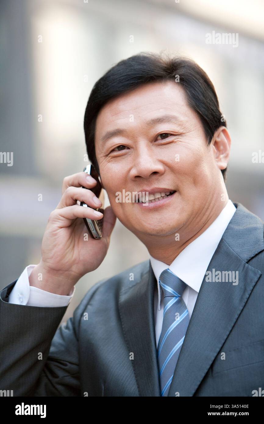 Ein chinesischer Geschäftsmann, der vor einem Stadtgebäude steht, plaudert auf seinem Handy. Die Geschäftsszene im Freien vermittelt professionelle Kommunikation und das städtische Arbeitsleben. Stockfoto